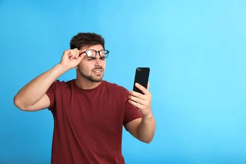 Young man with vision problems using smartphone on blue background, space for Stock Photos