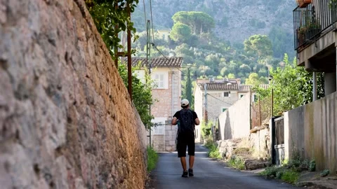 Young man walking with a backpack through a village towards green mountains.. Video stock 294178172