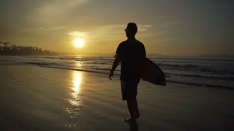 Young man walking on sandy beach holding longboard sunset rapid slow motion Video stock 84823786