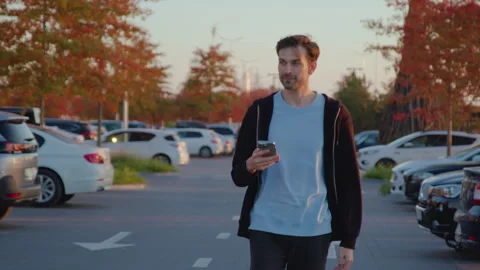 Young man walking through a car park, holding smartphone, surrounded by parked Video stock 315530753
