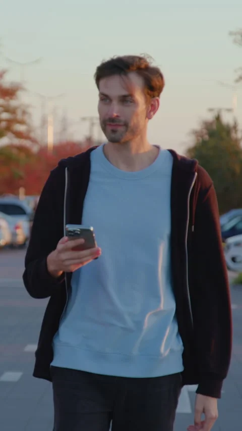 Young man walking through a car park, holding smartphone, surrounded by parked Stock Footage 318474833