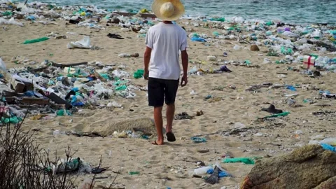 Young man walks on beach polluted by plastic junk looking at industrial Vídeos de archivo 151969981