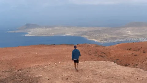 Young man walks to the edge of the cliff to observe the island of La Graciosa Stock Footage 136824923