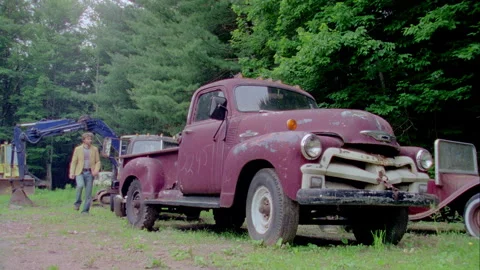 A young man walks by an old red truck in a junkyard Stock Footage 272269954