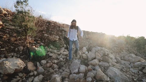 A young man walks over stones in the rays of sunlight with a tripod in his hands Stock Footage 105271853