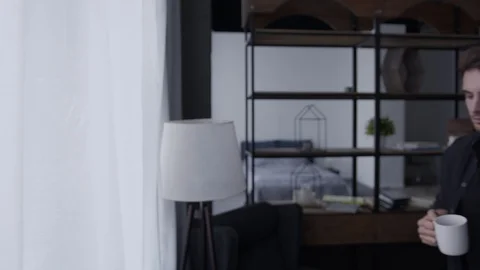 A young man walks to the window and looks into the distance, he also drinks 스톡 동영상 129330865