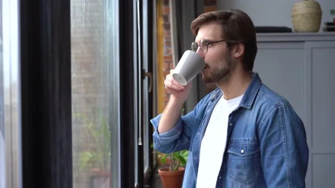 A young man walks up to the window with a mug of coffee or tea and takes a sip. Stock Footage 142876958