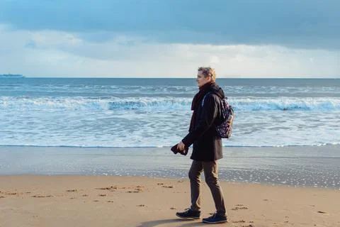 A young man in warm clothes with backpack walking on winter seaside and enjoying Stock Photos