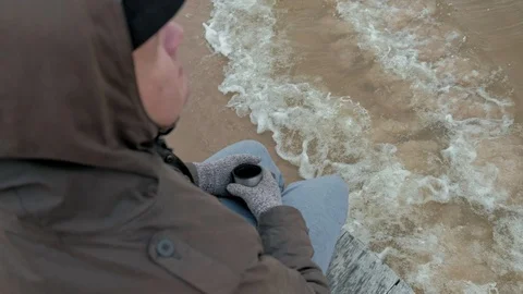 Young man in warm clothes sitting on the ocean shore, on a wooden coil, drinking Video stock 97256808