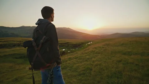 Young man watches the sunset while standing on a grassy hillside near a stream Stock Footage 345640627