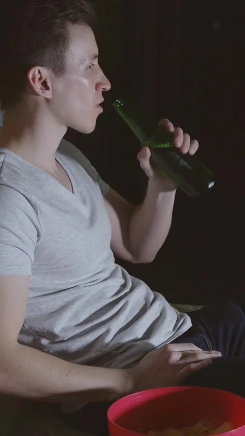 Young man watching tv at night drinking beer and eating chips from a bowl Stock Footage 344824370