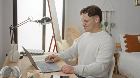 Young man waving hand at laptop on a bedroom desk inside an apartment build.. 動画素材 337424387