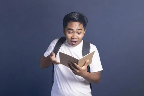 Young man wearing a backpack holding an open notebook with a shocked and be.. Stock Photos