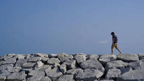 Young man wearing a cap walking down the rocky sand beach in Rayong, Thailand Stock Footage 156597152