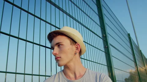 Young man wearing a hat, standing near a metal fence on a sunny day 스톡 동영상 306882012