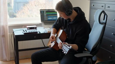 Young man wearing headphones creating music, playing an acoustic guitar while Stock Footage 322186331