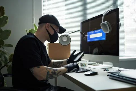 Young Man Wearing Mask Using Smartphone While Hacking Computer in Office 库存照片