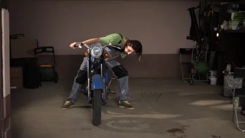 Young man wearing overalls sitting on a motorcycle in a garage, contemplating Stock Footage 340429230