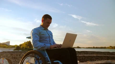 Young man in a wheelchair opens his laptop sitting in front of a beautiful b Stock Footage 202117006