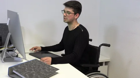 Young man in a wheelchair at a workplace at a computer Stock Footage 124899724