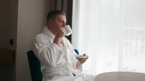 Young man in white bathrobe drinking coffee by window in hotel room Stock Footage 341950995