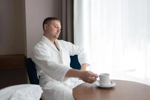 Young man in white bathrobe drinking coffee by window in hotel room Stock Photos