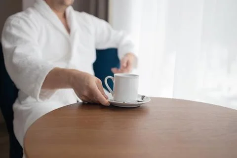 Young man in white bathrobe drinking coffee by window in hotel room Stock Photos