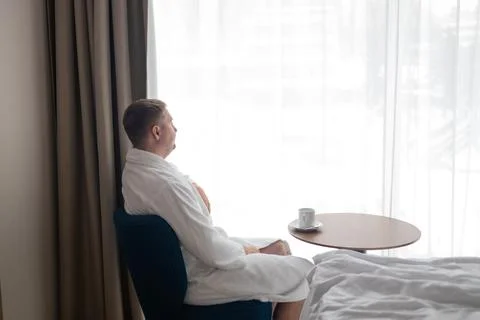 Young man in white bathrobe drinking coffee by window in hotel room Stock Photos