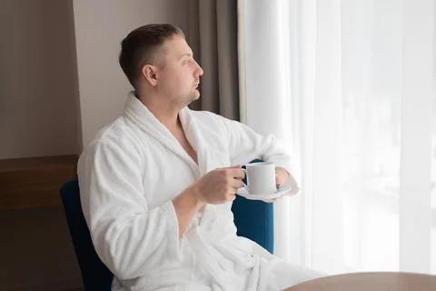 Young man in white bathrobe drinking coffee by window in hotel room Stock Photos
