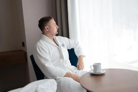 Young man in white bathrobe drinking coffee by window in hotel room Stock Photos