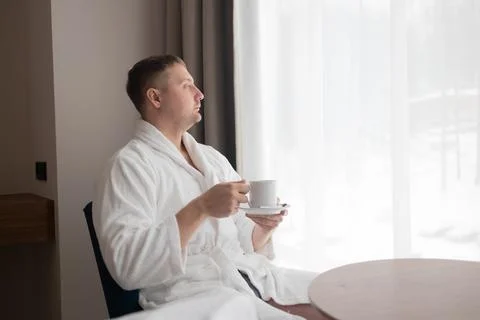 Young man in white bathrobe drinking coffee by window in hotel room Stock Photos