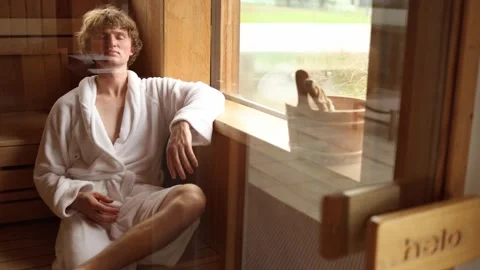 Young man in white bathrobe relaxing in sauna sitting near window with eyes clos Stock Footage 318778697
