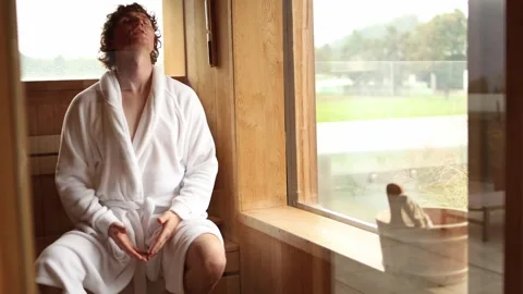 Young man in white bathrobe sitting in sauna breathing deeply and relaxing enjoy Stock Footage 318778703