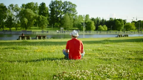 Young man in a white hat sits on the grass, looks at the pond, back view Stock Footage 139885767
