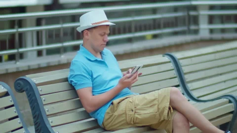 Young man in white hat sits on a city bench with a mobile phone in his hands Stock Footage 147696698
