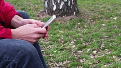 Young man with white phone sits in on street, in park, outdoor Stock Footage