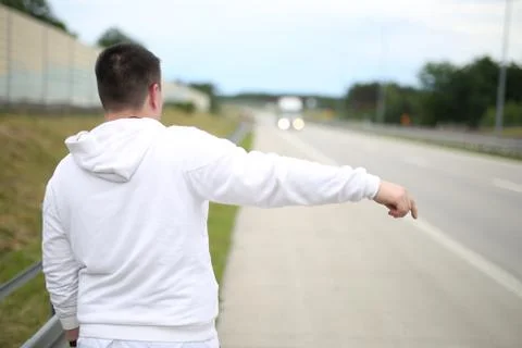Young man in a white pullover walking on the road and trying to stop a car. Stock Photos