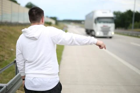 Young man in a white pullover walking on the road and trying to stop a car. Stock Photos
