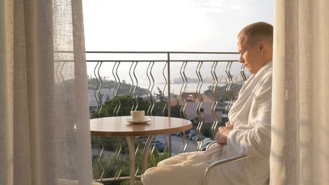 A young man in a white robe drinking coffee and thinking on the terrace. 4K Stock Footage 97964911