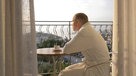 A young man in a white robe drinking coffee on the terrace. 4K Stock Footage 97965174