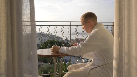 A young man in a white robe drinking coffee and using the phone on the terrace Stock Footage 97966218
