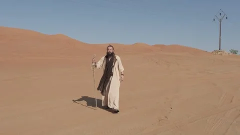 A young man in white shaman clothes with a staff in his hand walks on the sand Stock Footage 245876366
