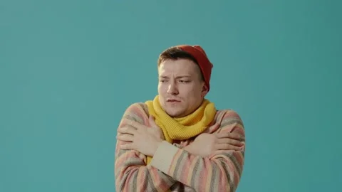 A young man in winter clothes on a blue background blows on hands, feels cold. Stock Footage 226219996