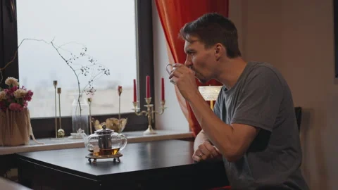 A young man on a winter day drinks tea by the window of the house Stock-Footage 294239745