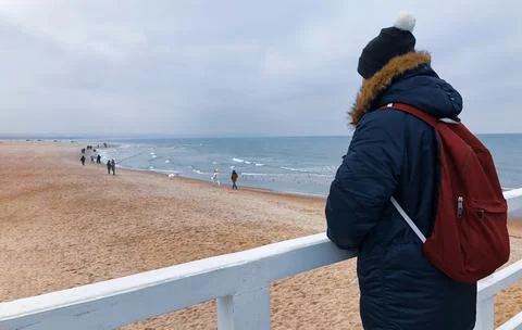 A young man in winter in a jacket, a hat and a backpack stands on the pier and Stock Photos