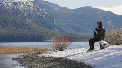 Young man in a winter jacket sits on a snowy log, pouring mate from a thermos. Stock Footage 327442565