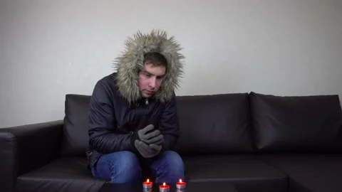 A young man in a winter jacket is warmed by candles. A man freezes at home Stock Footage 223752599