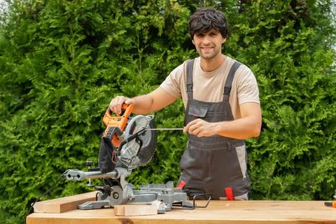 Young man in a work uniform repairs a miter saw using a screwdriver Stock Photos