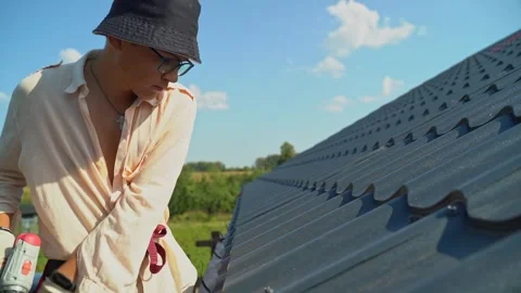A young man worker in a black Panama hat repairs a metal tile roof using an Stock Footage 265072957