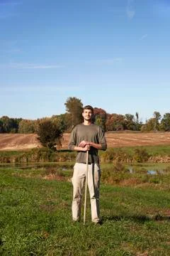 A young man in working clothes standing in a field, holding a tool. Stock Photos